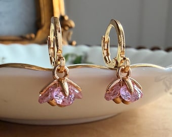 May include: A pair of gold-toned hoop earrings featuring dangling flower-shaped charms. Each charm is set with faceted light pink stones arranged to resemble petals, suspended from a small circular gold hoop closure.