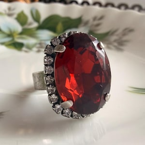 May include: A silver-toned ring featuring a large, oval-shaped, deep red faceted stone as the centrepiece. The central red stone is surrounded by a halo of small, clear, round-cut white stones set into the metal frame of the ring band.