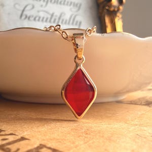 Peut inclure: Un collier doré avec un pendentif en forme de losange orné d'une pierre rouge translucide à facettes. Le pendentif est suspendu à une fine chaîne dorée, mettant en valeur une monture en métal poli qui encadre la pierre précieuse rouge.
