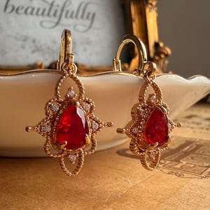 May include: A pair of gold-toned drop earrings featuring a central pear-shaped red gemstone surrounded by a decorative filigree frame accented with small clear stones. The earrings have a hinged lever-back closure and a textured metal finish.