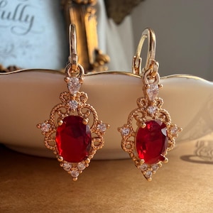 May include: A pair of gold-tone drop earrings featuring a central oval-cut red gemstone surrounded by intricate filigree metalwork and small clear accent stones. The earrings are suspended from a lever-back closure with a small clear teardrop stone.