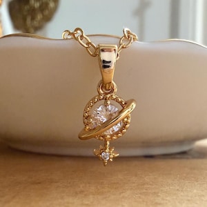 Puede incluir: Un colgante de collar en tono dorado con una piedra preciosa redonda y transparente en el centro, rodeada por un anillo que recuerda a un planeta. Un pequeño dije de estrella de cuatro puntas con una piedra clara cuelga debajo.