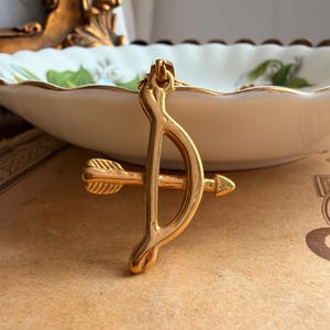 May include: A gold-toned metal pendant shaped like a bow and arrow. The bow is curved vertically, while the arrow is positioned horizontally across the centre of the bow. The piece has a smooth, polished finish and a small loop at the top for a chain.