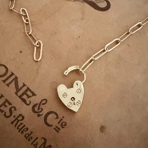 Puede incluir: Un collar dorado con un colgante de candado en forma de corazón. El candado está abierto, revelando los detalles del mecanismo de cierre. La cadena está hecha de eslabones alargados. El collar descansa sobre una superficie marrón con texto.