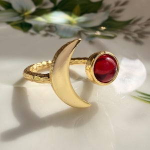 May include: A gold-toned open ring featuring a crescent moon shape on one end and a round, deep red cabochon stone on the other. The band of the ring has a textured, hammered finish, creating a distinct contrast with the smooth surface of the moon.
