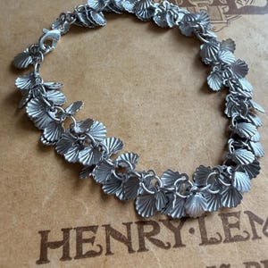 Op de afbeelding: Een zilverkleurige bedelarmband bestaande uit een reeks kleine, getextureerde schelpvormige bedels die in een doorlopend cirkelvormig patroon met elkaar zijn verbonden. De armband is voorzien van een karabijnsluiting en heeft een matte metalen afwerking.