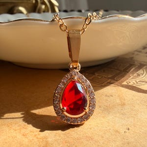 May include: A gold-toned necklace featuring a teardrop-shaped pendant with a vibrant red centre stone. The central red stone is surrounded by a halo of small, clear, sparkling stones set in a gold-toned metal frame, suspended from a matching chain.