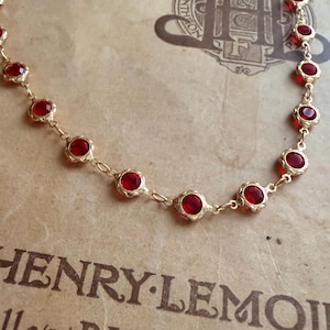 Puede incluir: Un collar dorado con una serie de piedras preciosas rojas. El collar descansa sobre un papel vintage con el texto "HENRY LEMOIN" y "PARIS-BRUXEL". Las piedras preciosas son redondas y están engastadas en un bisel decorativo.