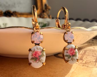 May include: A pair of gold-tone drop earrings featuring a round, pale pink crystal at the top and an oval, white ceramic cabochon at the bottom. The oval cabochons are decorated with a small, detailed pink rose floral pattern and green leaves.