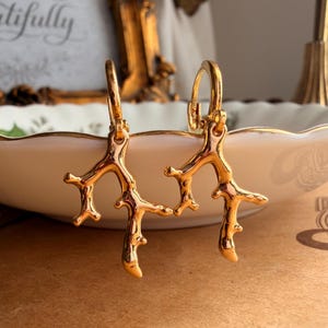 May include: A pair of gold-toned hoop earrings featuring dangling charms shaped like organic, branching coral structures. The earrings have a polished, reflective metallic finish and are suspended from circular hinged hoops.