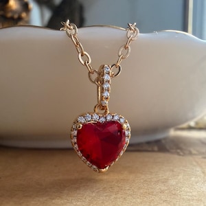 May include: A gold-toned necklace featuring a heart-shaped red pendant. The pendant is surrounded by a halo of small, clear, sparkling stones. The bail is also embellished with clear stones, and the chain consists of delicate, oval-shaped links.