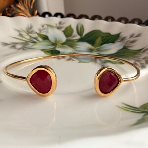 May include: A gold-toned open cuff bracelet featuring two teardrop-shaped, faceted deep red stones at each end. The metal band is thin and polished, creating a simple, continuous loop that remains open at the front to showcase the dark red gems.