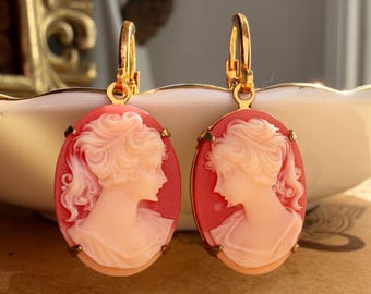 Può includere: Un paio di orecchini pendenti di forma ovale con uno sfondo color corallo e un ritratto di profilo in rilievo di colore chiaro. I cammei sono montati su cornici in metallo dorato e includono monachelle a leva color oro.