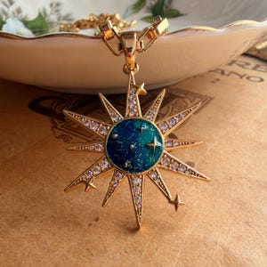 Könnte beinhalten: Ein goldfarbener Anhänger in Sternform mit einer zentralen, kreisförmigen blau-grünen Emaille-Einlage, die mit goldenem Glitzer gesprenkelt ist. Die Strahlen des Sterns sind mit klaren weißen Kristallen und kleinen sternförmigen Aussparungen verziert.