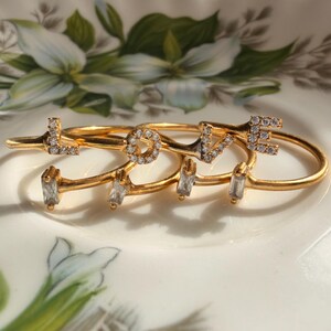 May include: A set of four gold-toned rings, each featuring a single letter from the word LOVE. The letters L, O, V, and E are embellished with small clear stones, while each ring band also includes a small rectangular clear stone accent.
