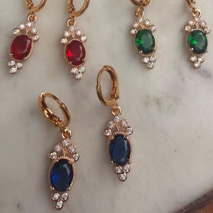 May include: Six gold-coloured hoop earrings with oval-shaped gemstones in red, green, and blue. Each earring has a decorative design with small, clear accent stones. The earrings are displayed on a light-coloured surface.