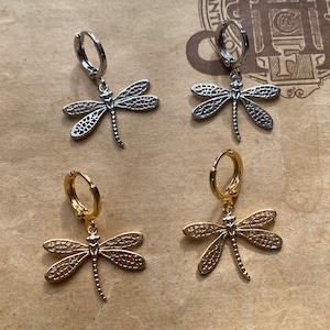 Op de afbeelding: Twee paar oorbellen met libellencharms. Het ene paar is zilver met een zilveren hoepel, het andere paar is goud met een gouden hoepel.