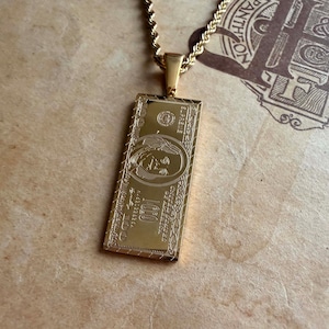 May include: Gold-toned rectangular pendant necklace featuring a detailed design of a $100 bill. The pendant hangs from a matching gold rope chain. The design includes the portrait of Benjamin Franklin and the text "100".