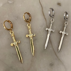 May include: Gold and silver sword earrings. Each earring has a detailed sword pendant suspended from a small hoop. The gold earrings have a shiny finish, while the silver earrings have a metallic appearance. The earrings are displayed on a marble surface.