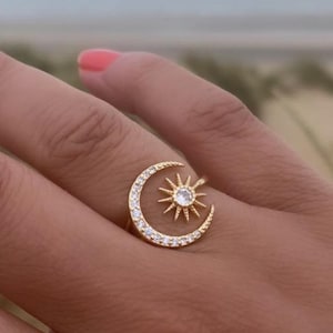 May include: Gold-toned ring featuring a crescent moon design encasing a sunburst. The moon is embellished with small, clear stones. The ring is worn on a finger with coral-colored nail polish. The background shows a beach scene.