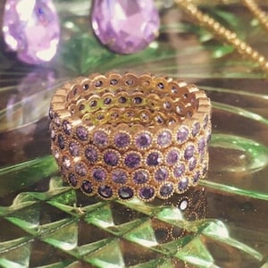 May include: A gold ring with multiple bands of small purple stones. The ring is on a green glass surface with two purple teardrop earrings and a gold chain necklace with a purple stone pendant.