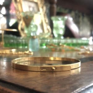 Könnte beinhalten: Ein goldfarbenes Armreif-Armband mit schlichtem, minimalistischem Design. Das Armband hat ein flaches Band mit einem kleinen, runden Verschluss.