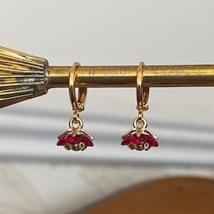 Op de afbeelding: Een paar gouden creolen met hangende rode bloemencharms. De oorbellen worden gepresenteerd op een gouden standaard.