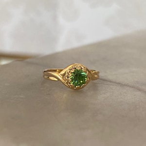 Könnte beinhalten: Ein goldener Ring mit einem runden, leuchtend grünen Edelstein in der Mitte. Der Ring hat ein detailliertes, verziertes Design um den Edelstein herum. Er liegt auf einer hellen Oberfläche mit unscharfem Hintergrund.