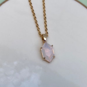 May include: A delicate gold chain necklace with a white gemstone pendant. The pendant is a geometric shape with multiple facets. The necklace is displayed on a white plate with a green and gold rim.