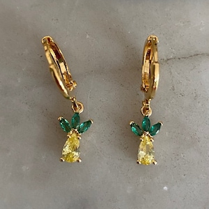 May include: A pair of gold hoop earrings with a dangling pineapple charm. The pineapple charm is made of yellow and green cubic zirconia stones.