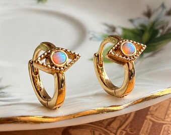 May include: A pair of gold hoop earrings featuring a central eye-shaped charm. Each charm is accented with a small, round, iridescent opal-like stone set in the middle of a textured, pointed metal frame that mimics an eye design.