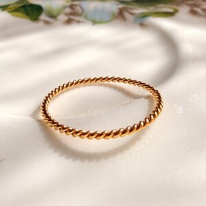 May include: A thin, gold-coloured ring featuring a continuous twisted rope texture. The band has a uniform, circular shape with a polished metallic finish that reflects light, creating a subtle shine along the spiraled surface of the metal.