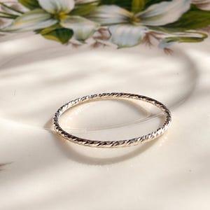 May include: A thin, circular silver ring featuring a twisted, textured surface that catches the light. The band has a continuous, repeating pattern of small, angled indentations along its entire circumference, creating a reflective, metallic finish.