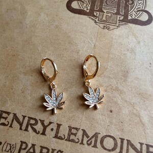 May include: A pair of gold-colored hoop earrings with a cannabis leaf charm. Each leaf is detailed with small, clear gemstones. The earrings are displayed on a textured, tan-colored surface with the text "HENRY LEMOINE (IX) PARIS-BRUXELLES".