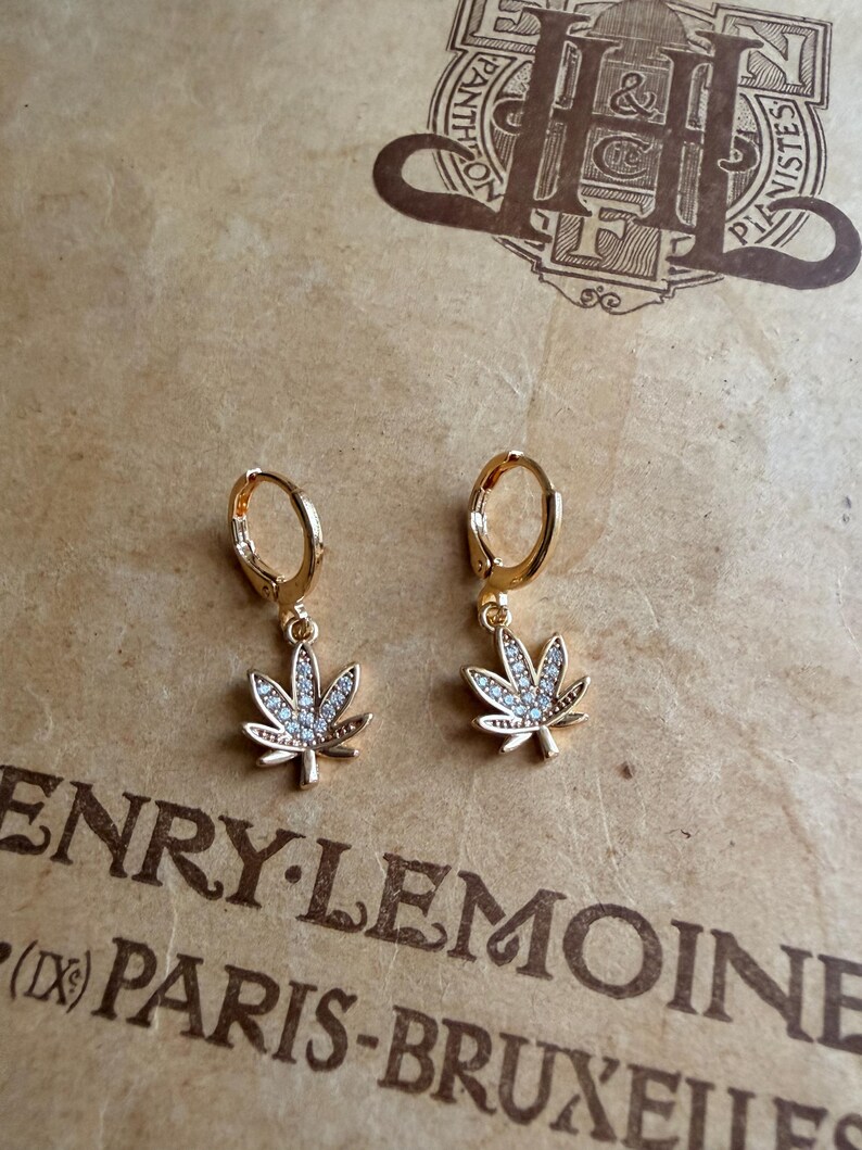 May include: A pair of gold-colored hoop earrings with a cannabis leaf charm. Each leaf is detailed with small, clear gemstones. The earrings are displayed on a textured, tan-colored surface with the text "HENRY LEMOINE (IX) PARIS-BRUXELLES".