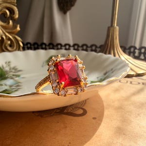 May include: A gold ring featuring a large rectangular red center stone surrounded by a halo of smaller clear stones. The band is textured with a fine pattern, and the setting includes additional baguette-cut clear stones along the sides.