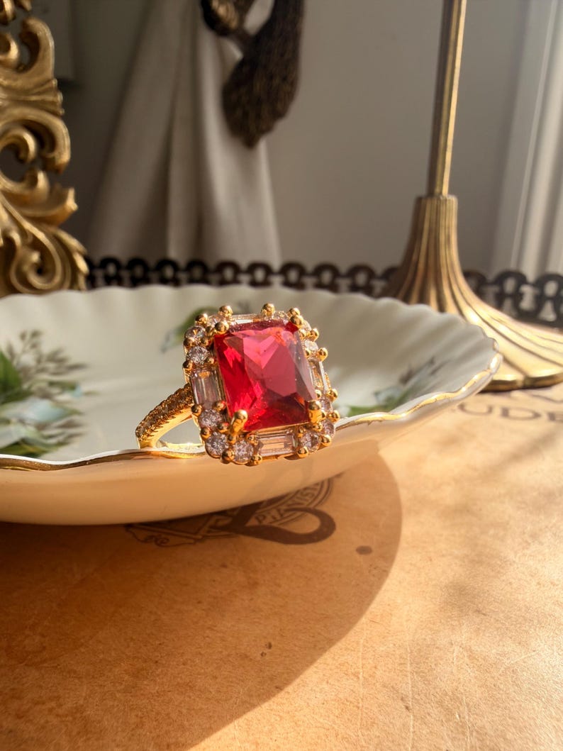 May include: A gold ring featuring a large rectangular red center stone surrounded by a halo of smaller clear stones. The band is textured with a fine pattern, and the setting includes additional baguette-cut clear stones along the sides.
