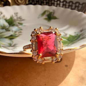 May include: A gold ring featuring a large rectangular red center stone surrounded by a halo of smaller clear stones. The band is thin and encrusted with tiny clear stones, and the setting is crafted with a metallic gold finish throughout.