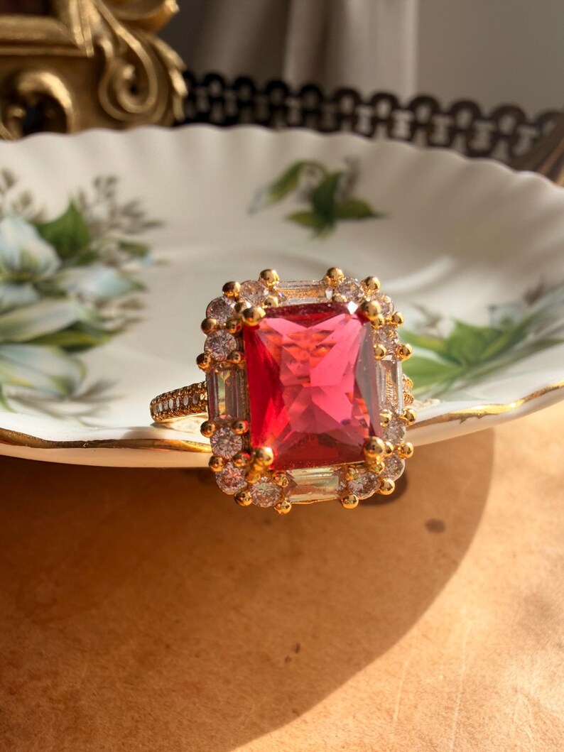 May include: A gold ring featuring a large rectangular red center stone surrounded by a halo of smaller clear stones. The band is thin and encrusted with tiny clear stones, and the setting is crafted with a metallic gold finish throughout.