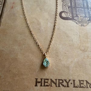May include: A gold-toned chain necklace featuring a single pear-shaped, light green gemstone pendant. The pendant is secured in a prong setting and hangs from a delicate, linked metal chain. The jewelry piece is displayed against a tan background.