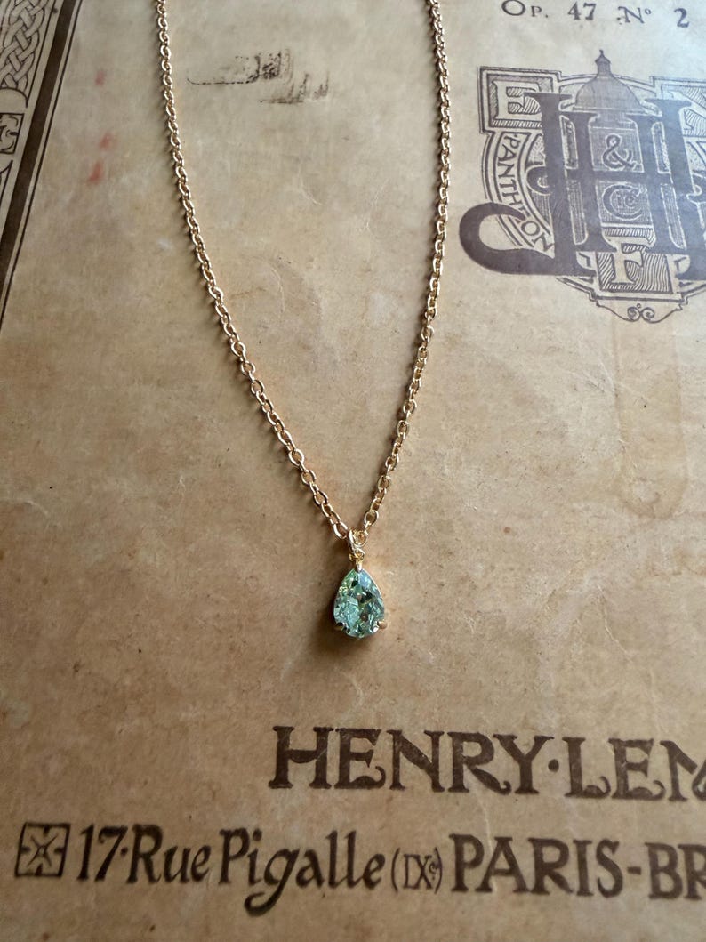 May include: A gold-toned chain necklace featuring a single pear-shaped, light green gemstone pendant. The pendant is secured in a prong setting and hangs from a delicate, linked metal chain. The jewelry piece is displayed against a tan background.
