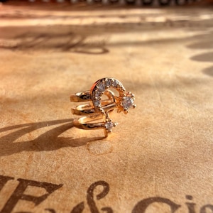 Könnte beinhalten: Ein goldfarbener Ohrklemmen-Schmuck mit einem Mond- und Stern-Design, verziert mit kleinen, klaren Edelsteinen. Der Schmuck hat drei Bänder und ist vor einem warmen, strukturierten Hintergrund platziert.