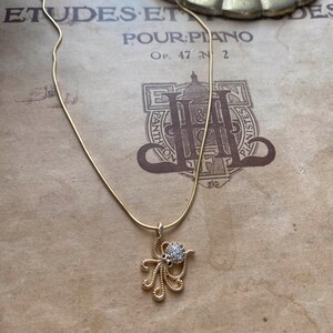 May include: A gold-colored necklace with a pendant shaped like an octopus. The octopus pendant has a round, gem-studded head and delicate, swirling tentacles. The necklace is displayed on a vintage sheet music background.