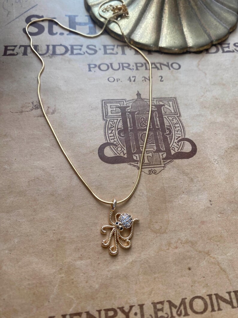 May include: A gold-colored necklace with a pendant shaped like an octopus. The octopus pendant has a round, gem-studded head and delicate, swirling tentacles. The necklace is displayed on a vintage sheet music background.