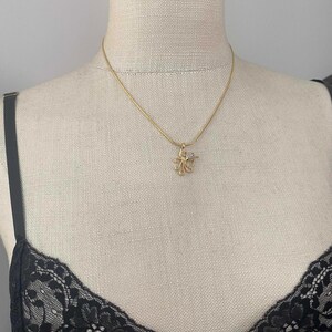 May include: A gold-colored necklace with a small, flower-shaped pendant. The pendant has a central clear stone and is suspended from a delicate chain. The necklace is displayed on a mannequin.