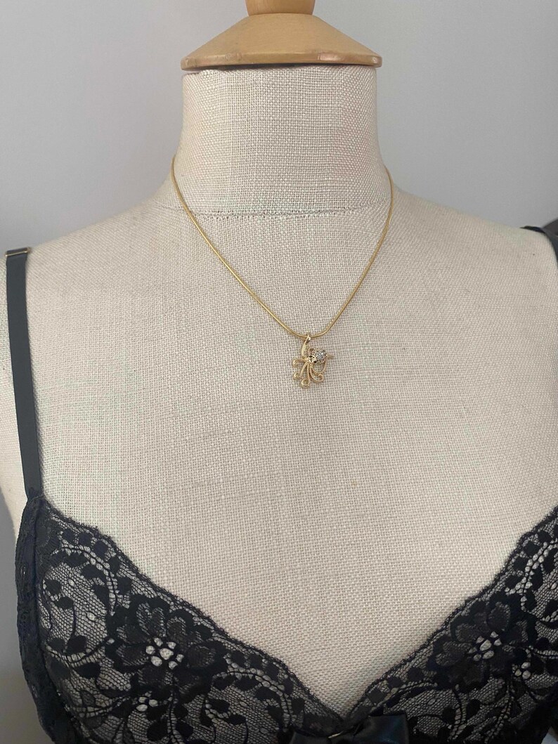 May include: A gold-colored necklace with a small, flower-shaped pendant. The pendant has a central clear stone and is suspended from a delicate chain. The necklace is displayed on a mannequin.