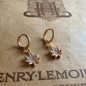 May include: A pair of gold-colored hoop earrings. Each earring features a small, detailed cannabis leaf pendant adorned with clear crystals. The earrings are displayed on a vintage paper background with text.