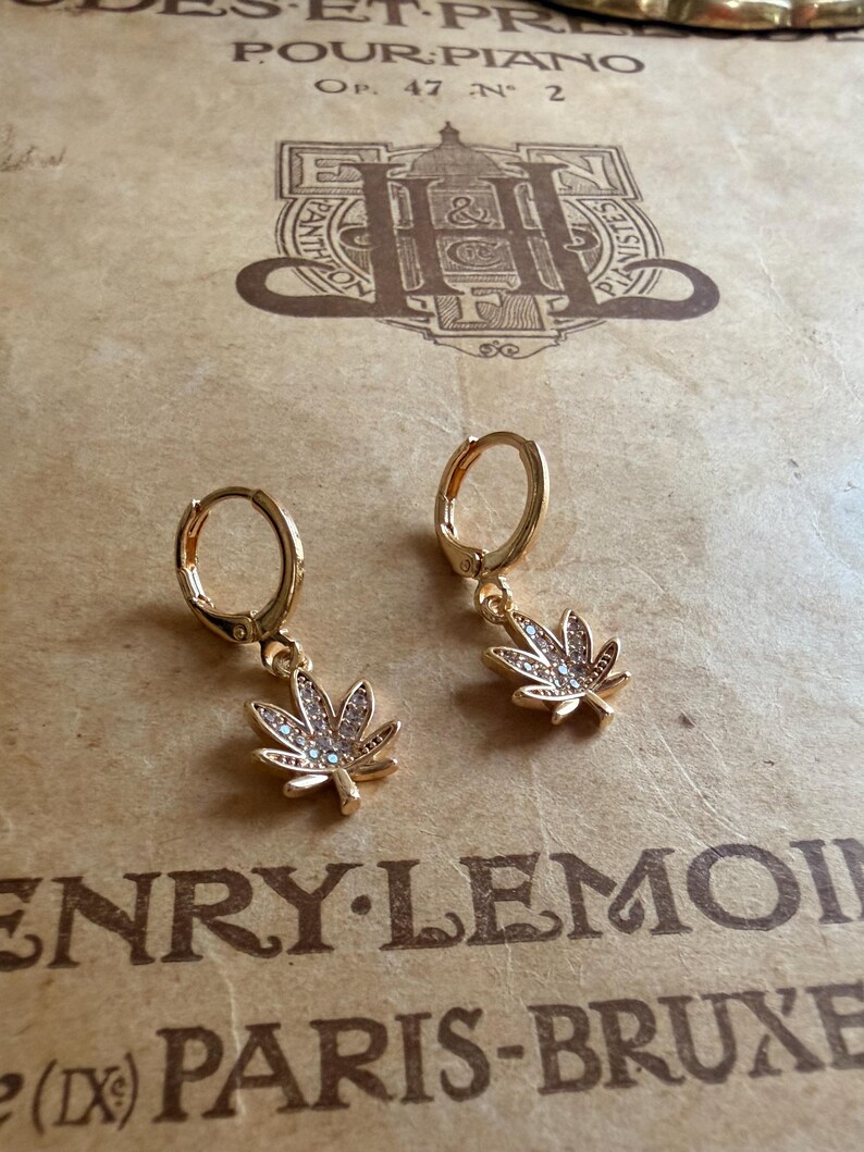 May include: A pair of gold-colored hoop earrings. Each earring features a small, detailed cannabis leaf pendant adorned with clear crystals. The earrings are displayed on a vintage paper background with text.