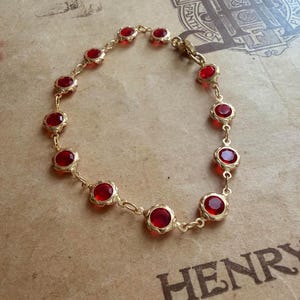 Könnte beinhalten: Ein goldfarbenes Armband mit einer Reihe von runden, tiefroten Edelsteinen. Die Edelsteine sind in verzierten, geschwungenen Fassungen gefasst und durch zarte Goldketten verbunden. Das Armband wird auf einem Vintage-Papierhintergrund präsentiert.