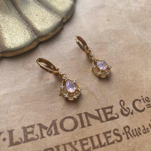 May include: A pair of gold hoop earrings with a teardrop-shaped pink stone surrounded by smaller clear stones.